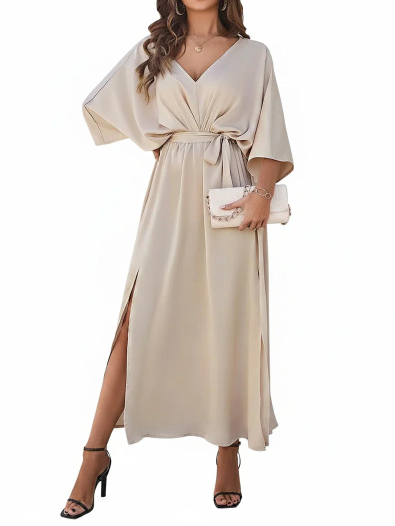 Beige maxi jurk met diepe V-hals, vleermuismouwen en strikceintuur gedragen door model met hakken