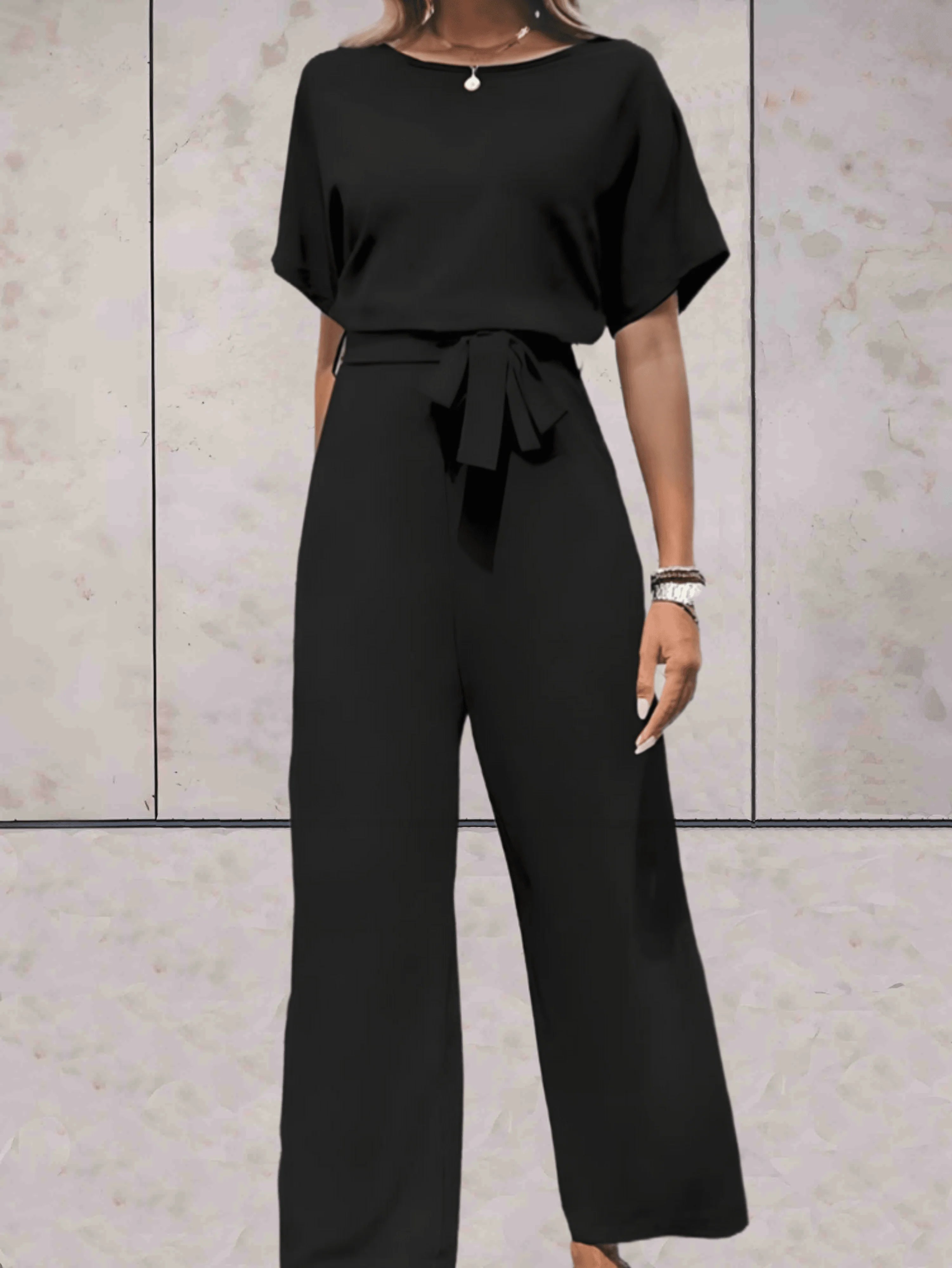 Zwarte jumpsuit met korte mouwen en strikceintuur gedragen door model voor lichte muur