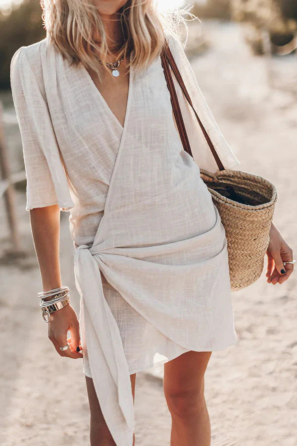 Beige wikkel mini jurk met asymmetrische zoom en korte mouwen, gedragen op het strand met rieten tas
