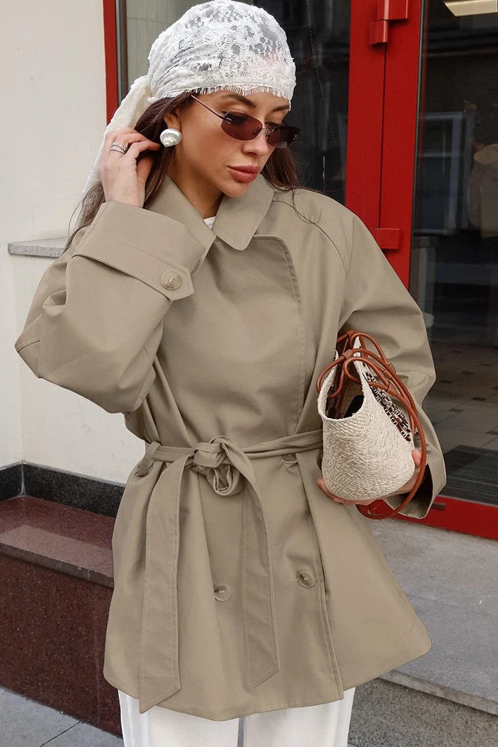 Dames korte trenchcoat in beige met ceintuur en klassieke revers, gedragen met witte broek