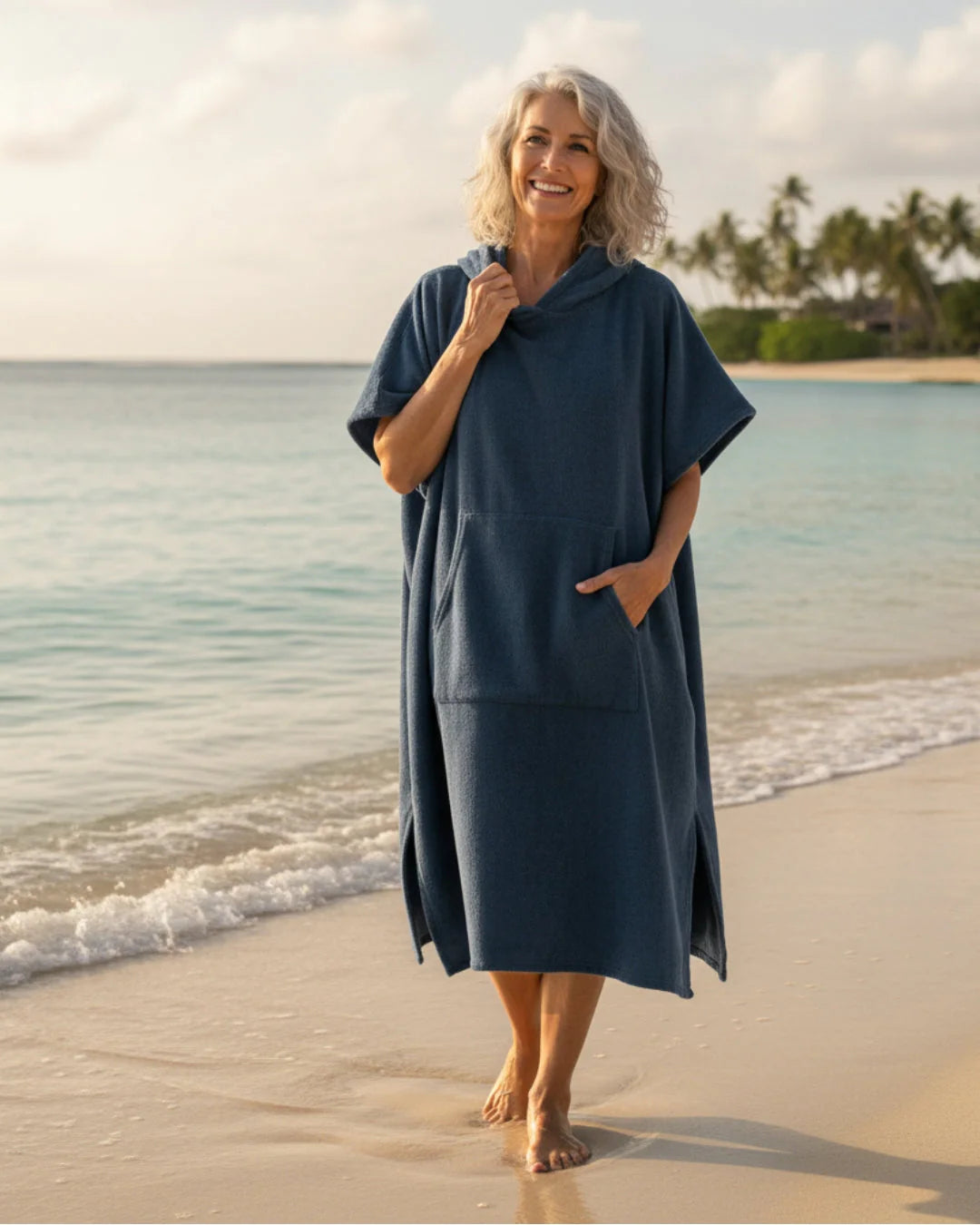 Vrouw draagt donkerblauwe strand poncho jurk met capuchon en zakken op het strand bij zonsondergang