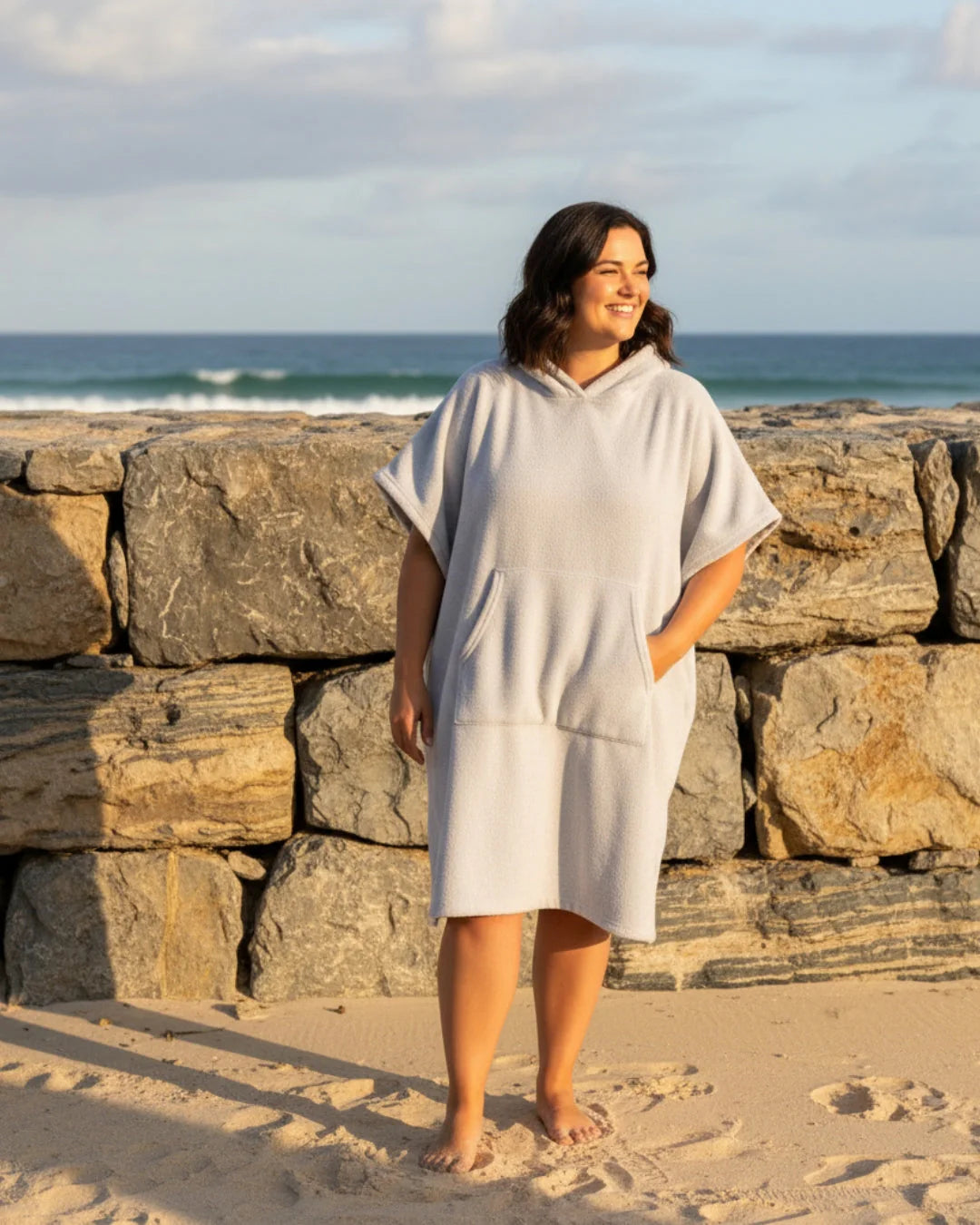 Vrouw loopt op strand in oversized poncho jurk met zijsplitjes en ontspannen silhouet